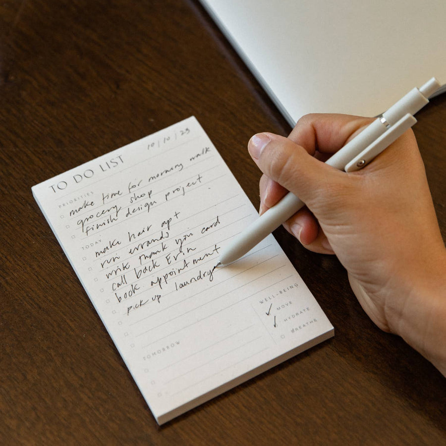 Hand writing on a to-do list with a pen, next to an open laptop on a wooden surface.
